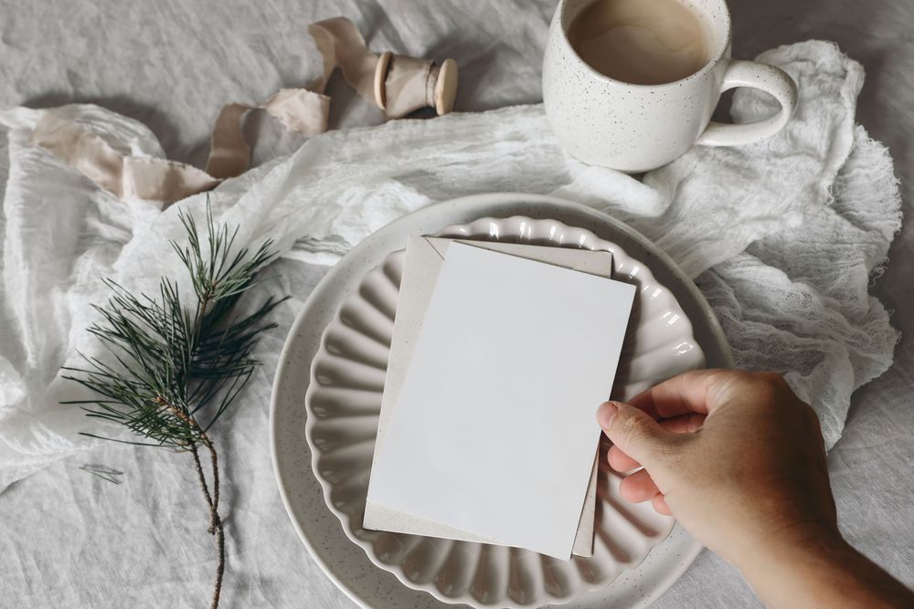 Hand placing blank card, invitation mockup on ceramic plate with pine branch on tablecloth