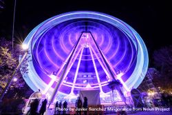 Slow shutter shot of purple lit ferris wheel at night with people queuing in front 5lL9o5