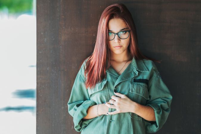 Stylish confident bright woman in glasses leaning on wall and looking at camera