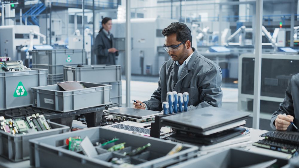 Indian And Caucasian Male Workers Taking Apart Laptops To Recycle Components For Production Of Microchips At Automated Electronics Factory With Robotic Arms.