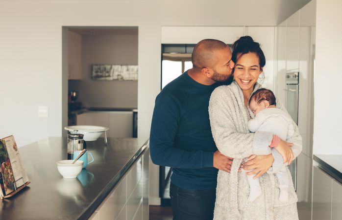 Man kissing his wife holding a newborn baby boy in kitchen