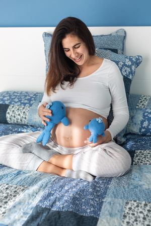 Tender pregnant woman sitting on bed with soft toys