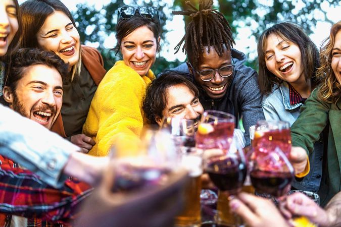 Multi-ethnic group of excited people having fun toasting wine and beers together at terrace birthday party