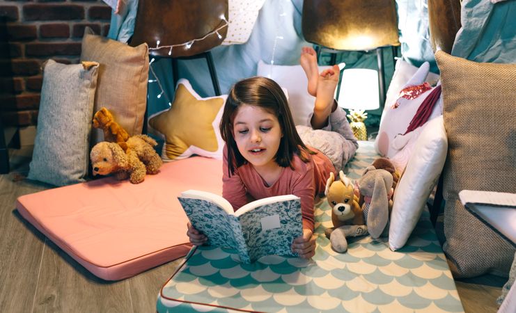Happy little girl reading and relaxing lying over sleeping mat in a cozy tent at home