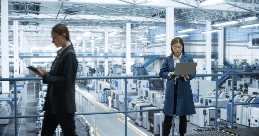 Thoughtful Asian Engineer Monitoring and Analyzing Work at a Modern Electronics Factory with Automated Robot Hands Making Devices. Female Work Colleague Passing By with a Tablet Device