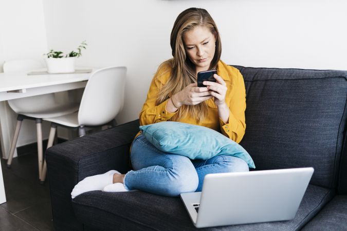 Long haired female sitting on sofa with laptop computer working remotely from home checking phone