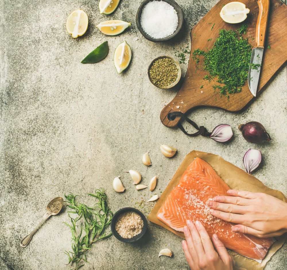 Hand rubbing salt on salmon fillet with fresh herbs, limes, onions, bread board, hand, copy space