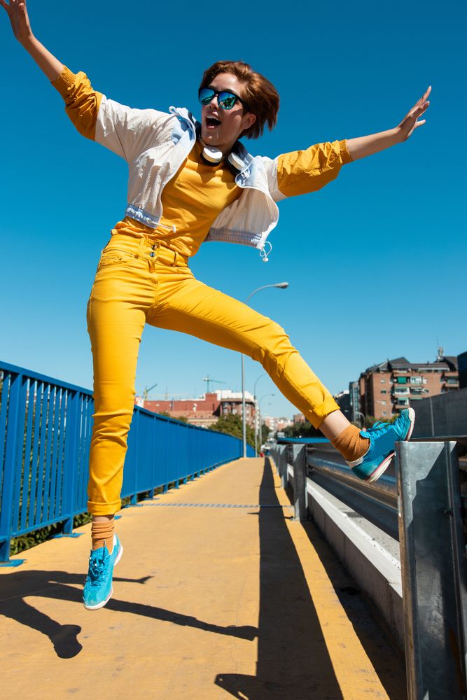 Stylish young carefree woman in vibrant clothes having fun on street bridge against blue sky