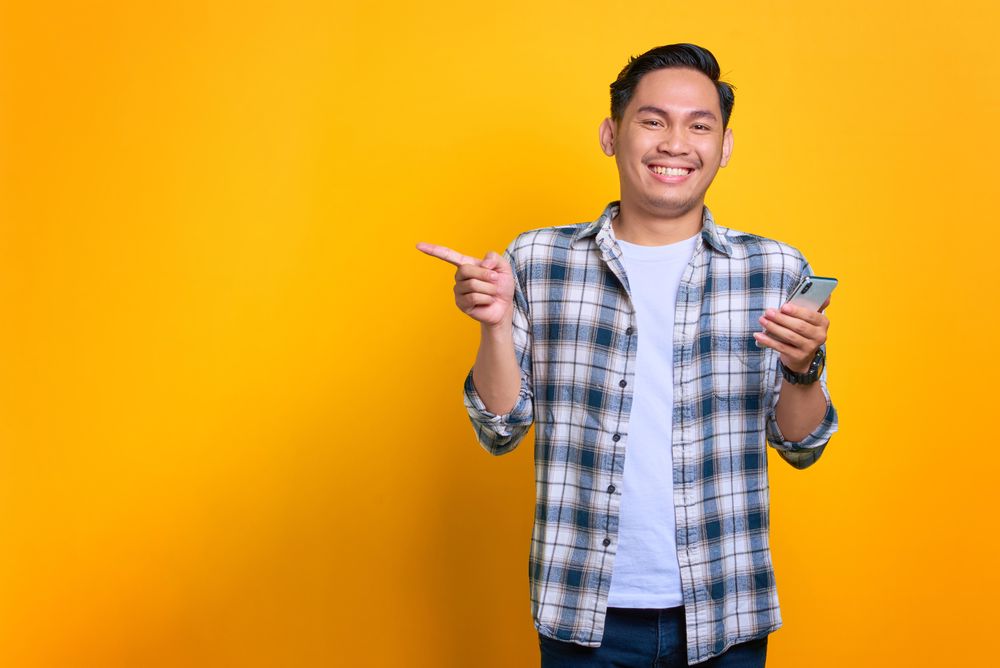 Smiling Asian male holding his phone and pointing to left