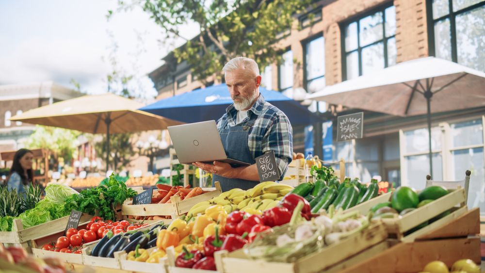 Portrait of a Street Vendor Working on Laptop Computer while Standing in a Farmers Market Stall with Fresh Natural Agricultural Products.
