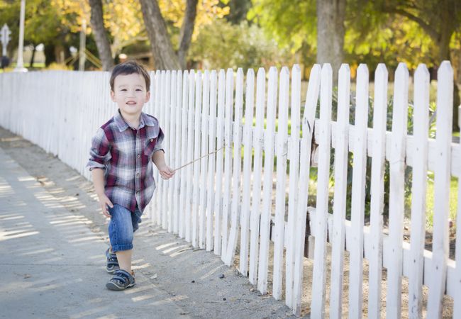 Young Boy Walking with Stick Along White Fence