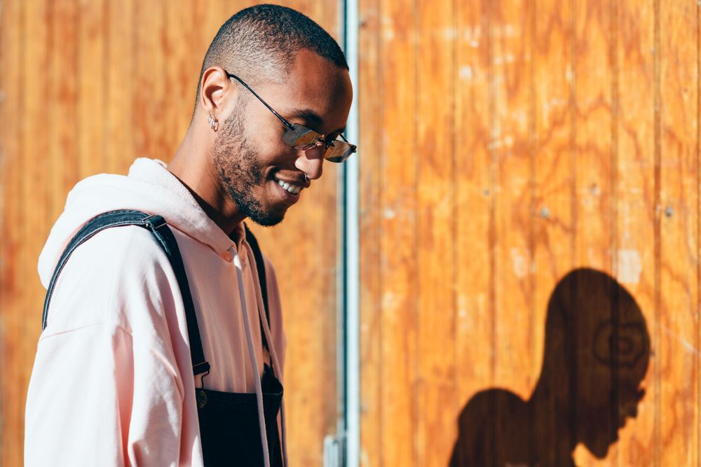 Male wearing overalls and smiling and looking down standing by a wooden wall