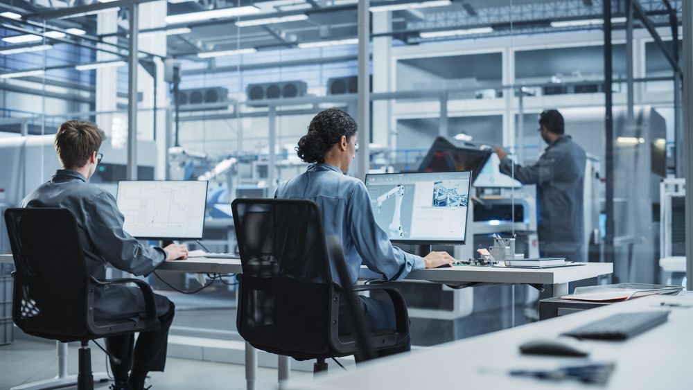 White Male Programmer And Hispanic Female Automation Engineer Using Desktop Computers At Modern Factory With Robotic Arms.