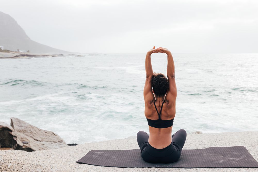 Female sitting next to the ocean sitting crossed legged stretching arms up