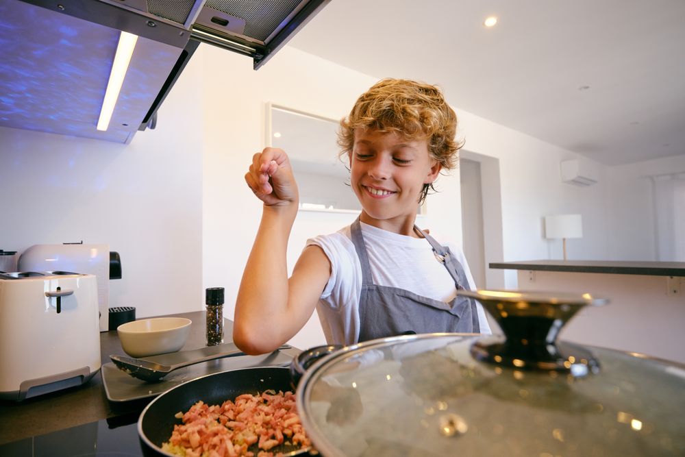Satisfied boy salting bacon in frying pan while cooking