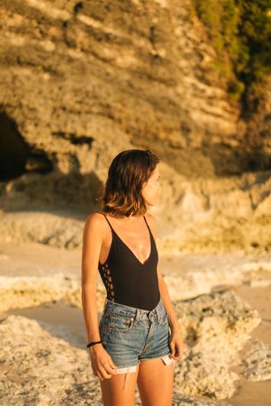 Woman in bathing suit and denim jeans looking to her side standing in front of cliff