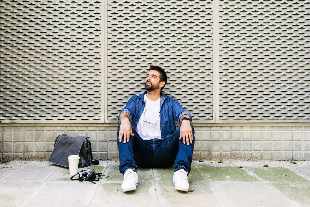 Latino man in denim sitting on pavement outside with cup of coffee