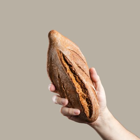 Hand holding a loaf of rustic whole wheat bread on a beige background.