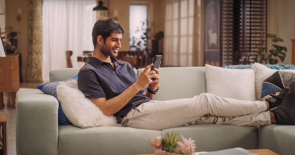 Indian Man Lying on Sofa Using Smartphone, Exploring Online Shopping,