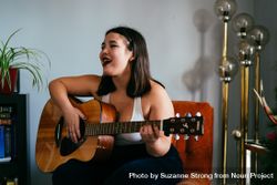 Young woman singing and playing guitar in house sitting on orange chair 4d8YA4
