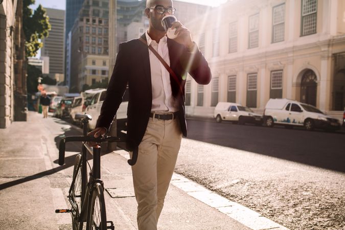 Man in sports jacket walking with cycle and drinking coffee on city street