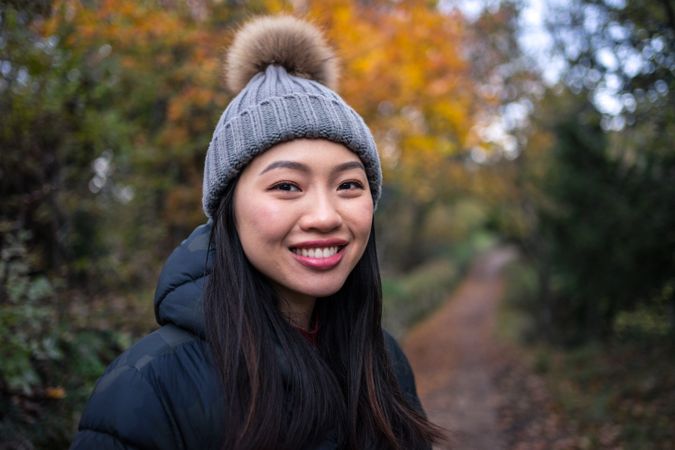 Smiling Asian woman in warm clothes standing in park with trees and stony fence