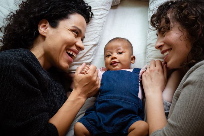 Two laughing women lying on a bed with baby girl while holding her hands