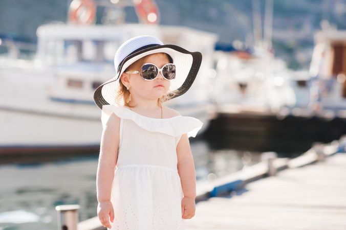 Young girl in light dress with hat and sunglasses standing beside docked boats