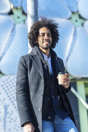 Portrait of happy Black man in jacket with warm beverage standing outside leaning on pole