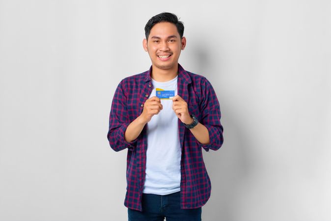 Happy Asian male in grey studio holding up credit card with his hands