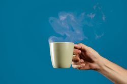 Hand holding a steaming hot coffee mug on a solid blue background.