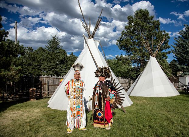 Two Native American men in full traditional regalia standing outside with teepees