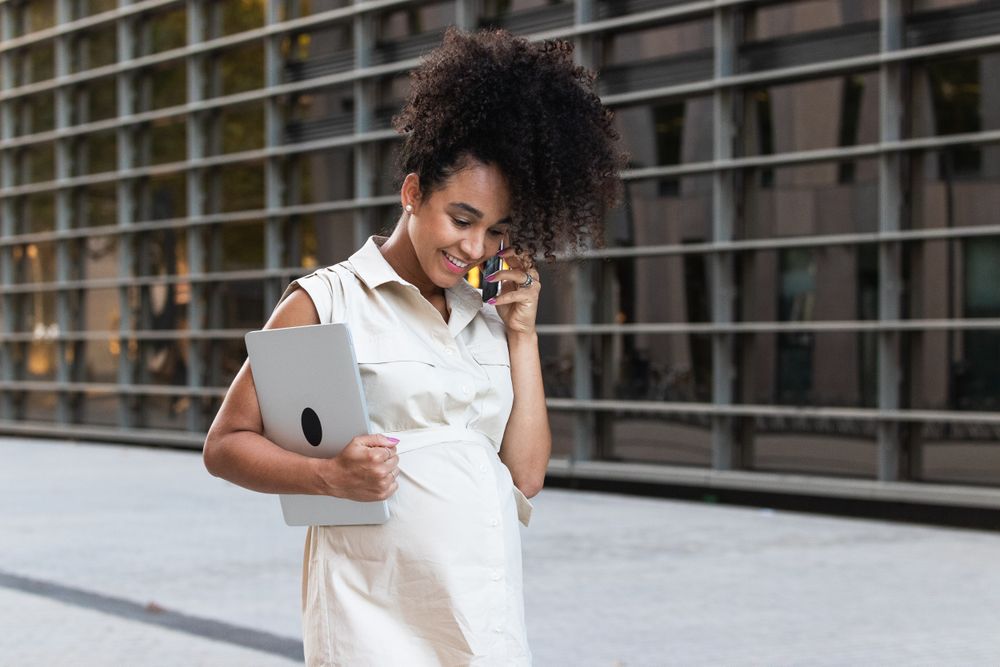 Smiling Black pregnant woman talking on smartphone and carrying a laptop