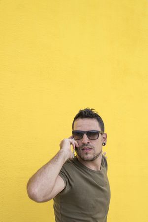 Serious male leaning on yellow wall talking on cell phone