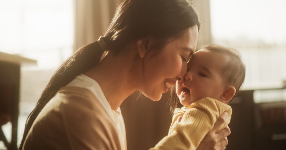 Motherly Affection Concept: Close Up Portrait of an Affectionate Asian Mother Touching Noses with her Baby in a Cosy Naturally Lit Bedroom.