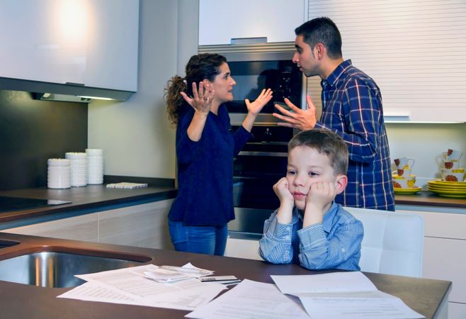Frustrated couple arguing in front of son with bills strewn on the kitchen counter