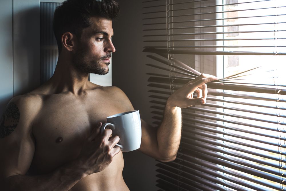 Man standing by a windows blind with a cup of coffee