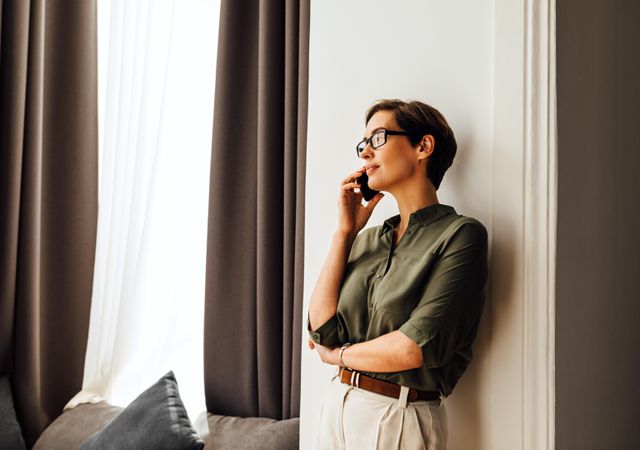 Serious woman leaning on wall next to a window taking a phone call