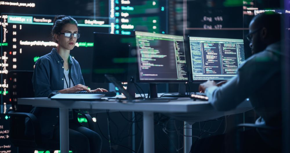 Diverse Young Man and Woman Working on Desktop Computers, Surrounded by Big Screens Displaying Code Prompts.