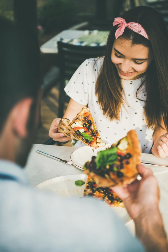 Couple eating pizza al fresco with wine, woman looking down smiling
