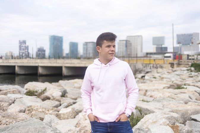 Male teenager standing against cityscape with hands in pocket on coastal breakwaters while looking away
