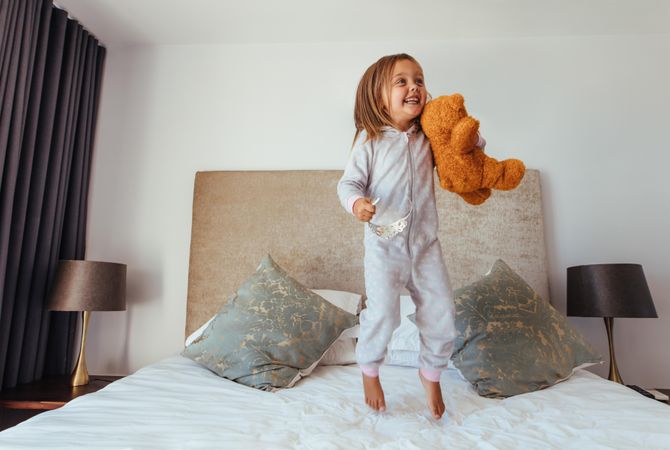 Joyful little girl jumping on bed with teddy bear