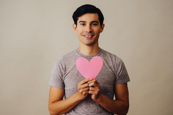 Romantic happy Hispanic male holding cut out pink heart to his chest