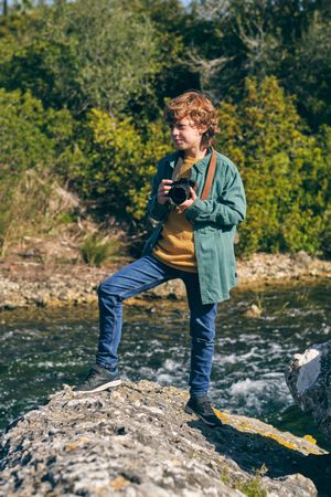 Content boy with photo camera standing on stone near brook