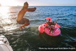 Playful male jumping midair into ocean water with friends floating on toy nearby 5rGLp4
