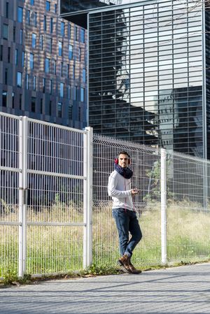 Male in headphones holding smartphone and looking around while leaning on a metallic fence outside