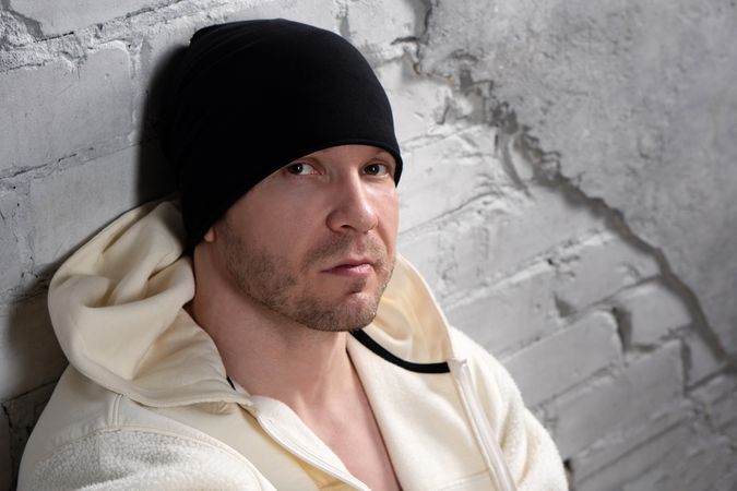 Man in beanie and fleece hoodie sitting against wall.