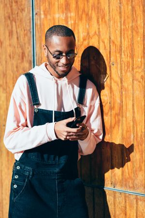 Male wearing overalls and smiling and checking phone standing by a wooden wall, vertical