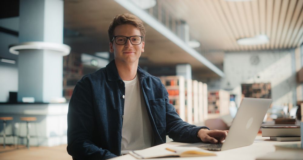 Confident Young Male Student Working On a Laptop in a Modern Library