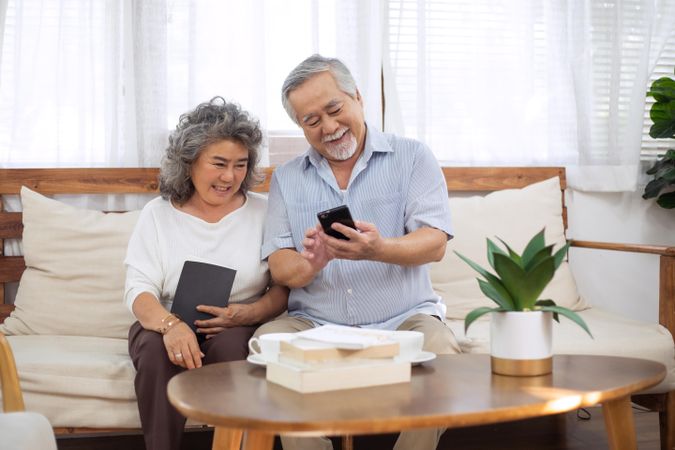 Happy Asian mature couple using smartphone together on couch in living room